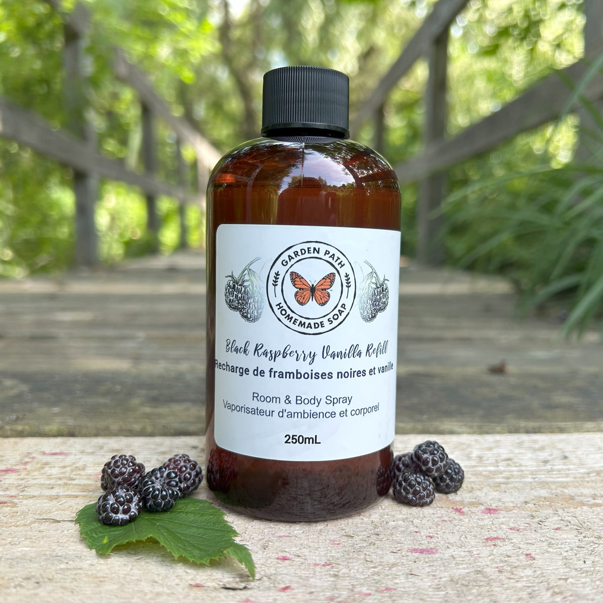 Black Raspberry Vanilla Room and Body spray refill bottle 250ML by Garden Path Homemade Soap on a wooden path with black raspberries.