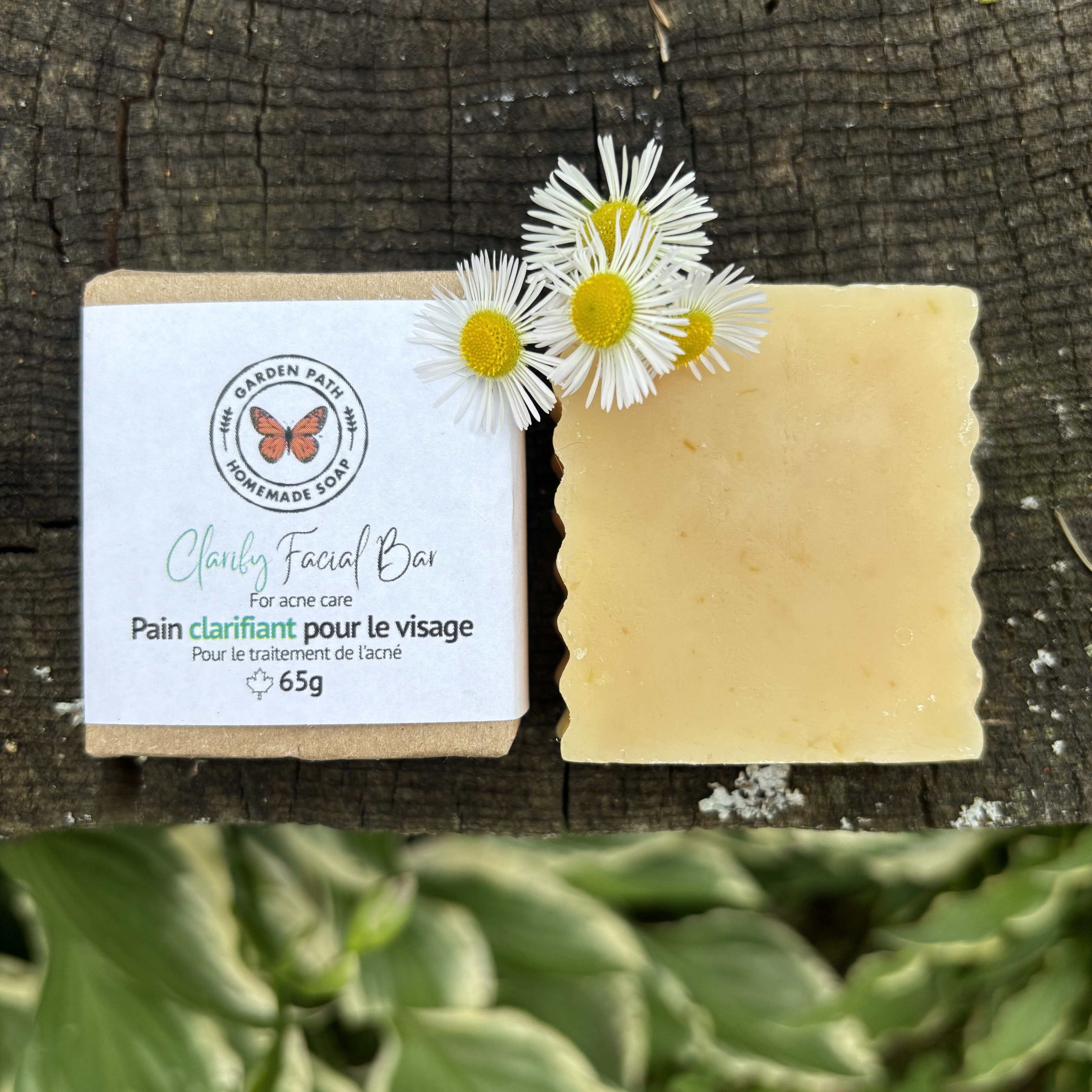 A handmade facial bar soap Clarify Facial Bar for acne care shown next to its paper packaging, placed on a rustic dark wood surface with white flowers on top. 100% Natural Ingredients - Garden Path Homemade Soap