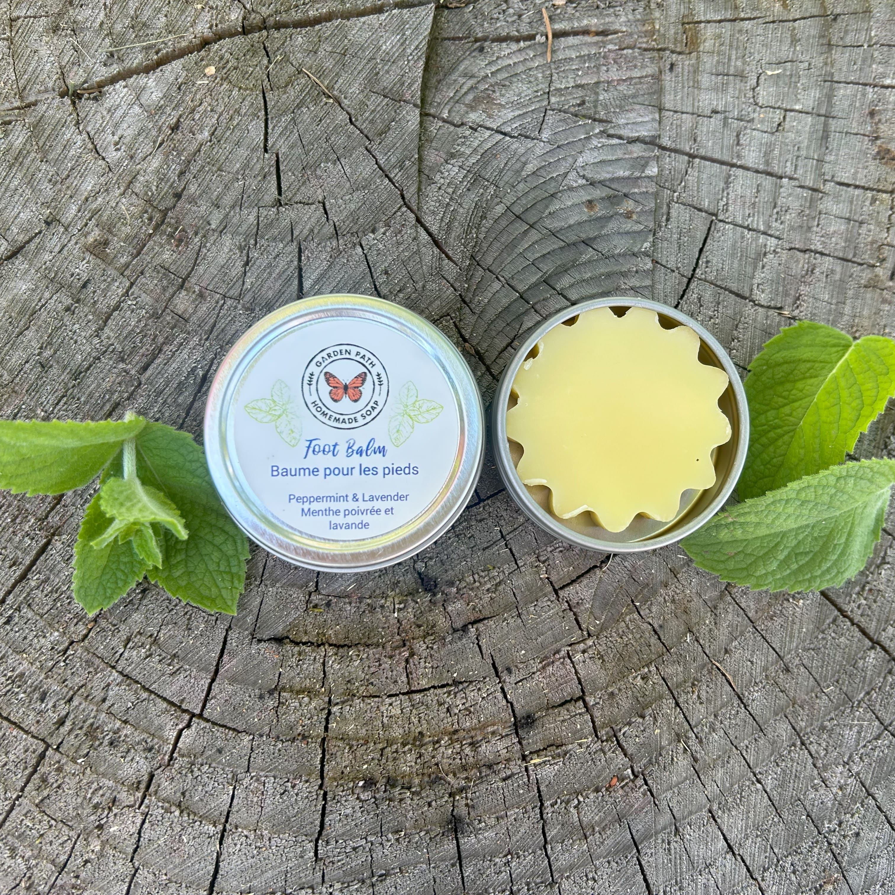 Tin of foot balm with a lid on a wooden surface with green mint leaves beside by Garden Path Homemade Soap. 100% Natural Ingredients | Intense Moisturizer