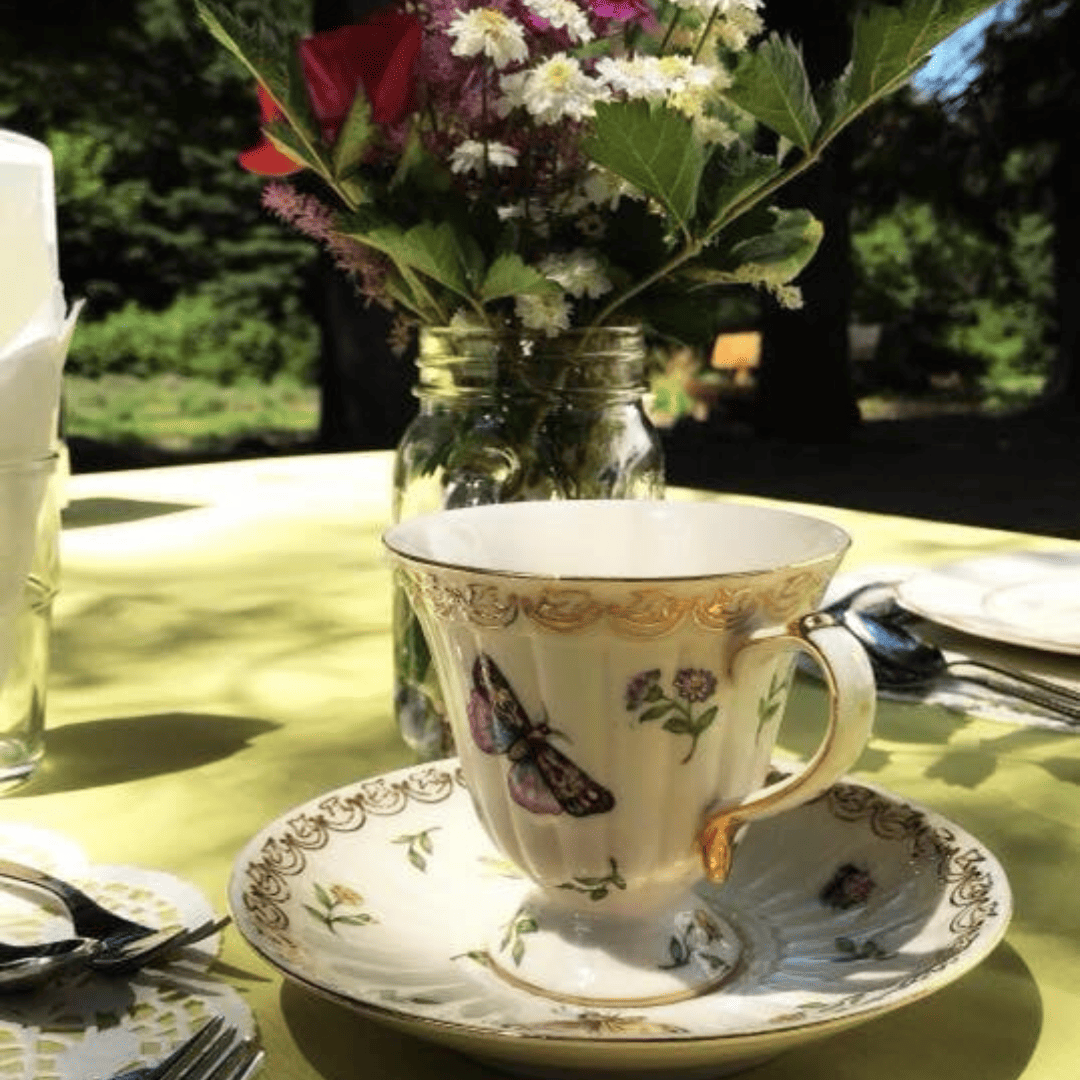 Decorative teacup with butterfly design on a saucer, placed on a table outdoors with a vase of flowers.