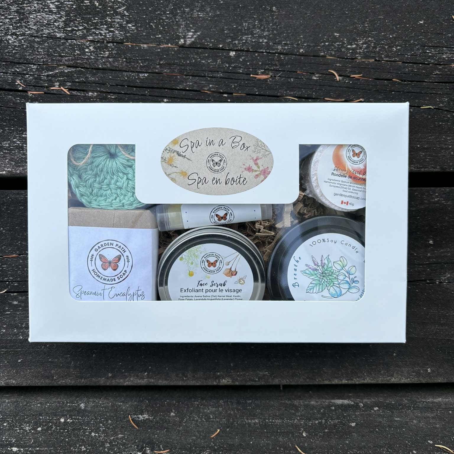 Spa in a Box set with various products like bar soap, facial scrub, small candle, shower puck and lip balm on a wooden surface by Garden Path Homemade Soap