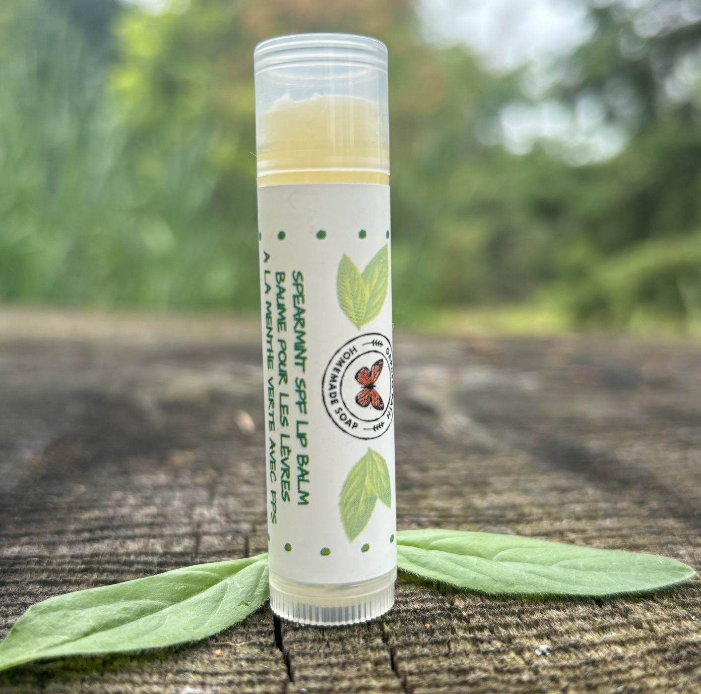 A stick of Spearmint SPF Lip Balm placed on a wooden surface with leaves in the background by Garden Path Homemade Soap. 100% Natural Ingredients