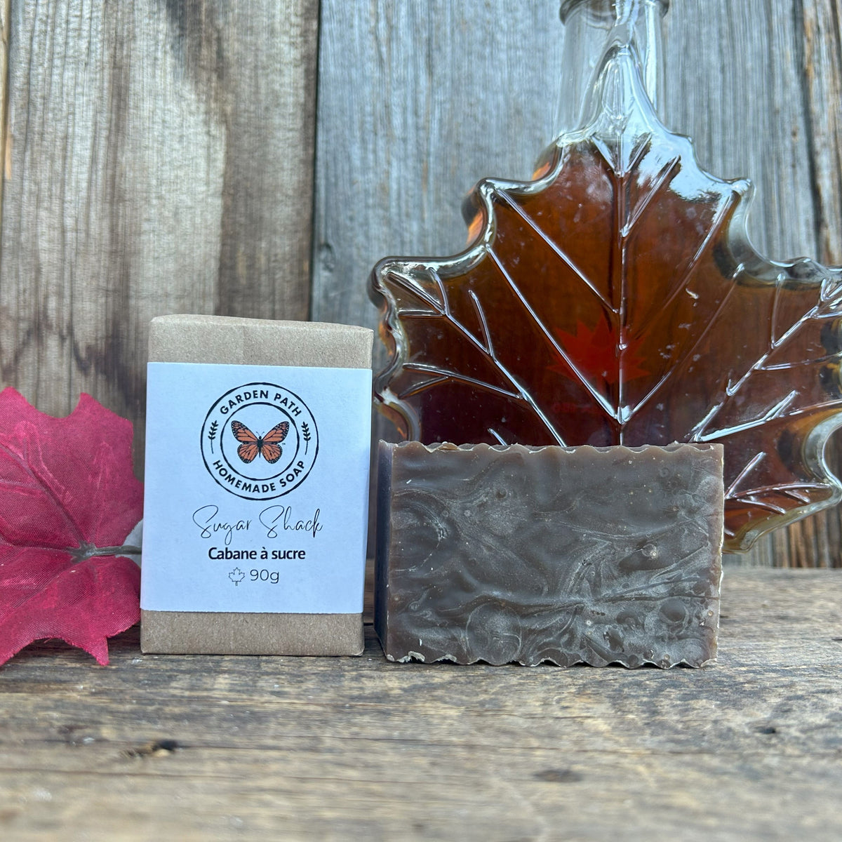 Garden Path Homemade Soap's Sugar Shack Bar of soap with packaging and a maple leaf glass container filled with maple syrup on a wooden surface. Lightly Scented Maple Fragrance & Natural Maple Sugar Sand