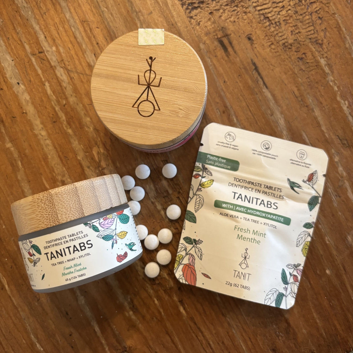 Three TANNTABS oral care products arranged on a wooden tabletop: a round bamboo-lidded jar, a stand-up pouch, and several white tablets scattered between them. The products have floral labels and the brand name TANNTABS is clearly visible.