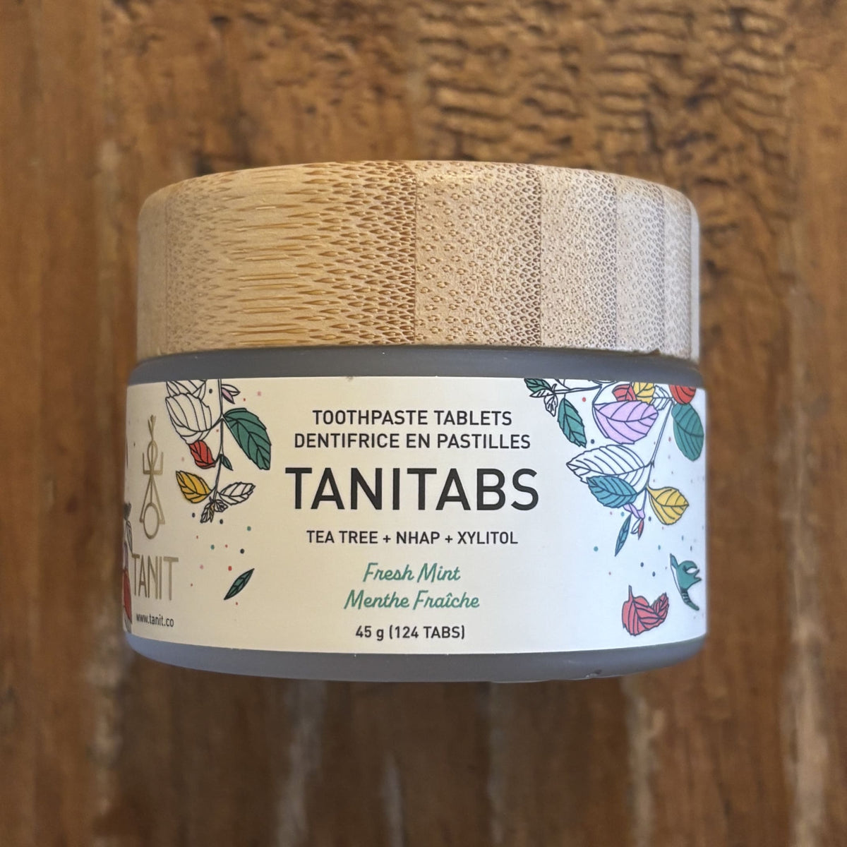 A jar of TANITABS toothpaste tablets in Fresh Mint flavor, featuring a light wood lid and a white label with botanical illustrations, set against a rustic wooden background.