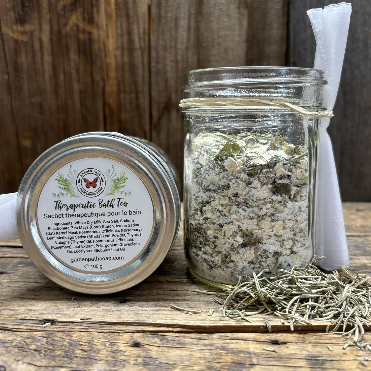Glass jar and tin labeled 'Therapeutic Bath Tea' on a wooden surface with herbs.