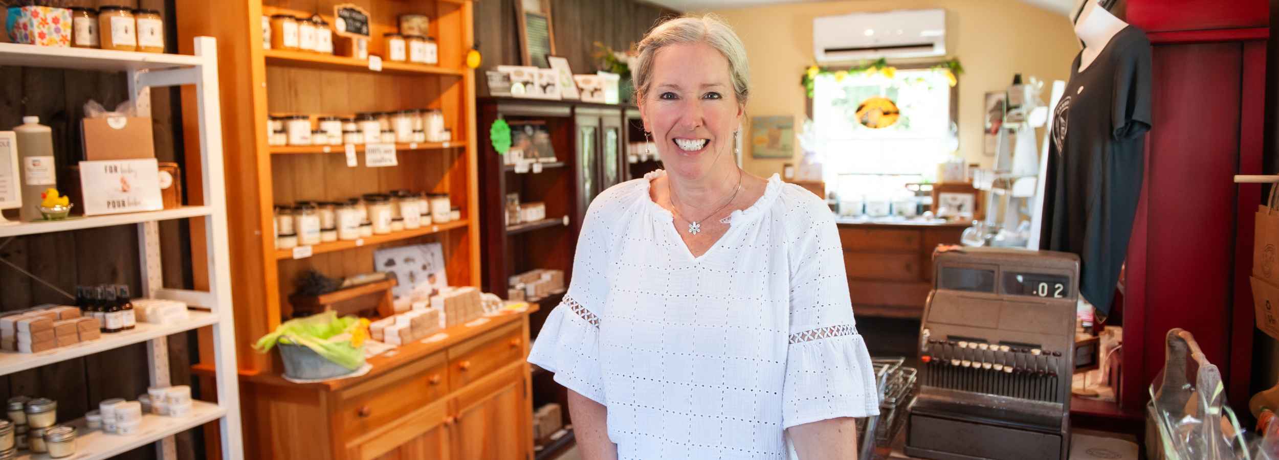 Tara, CEO of Garden Path Homemade Soap, stands in a retail shop interior wearing a white eyelet blouse. Shelves with jars and all natural products, a cash register, and store fixtures are visible around her.