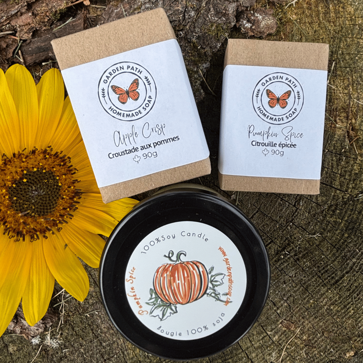 Apple Crisp and Pumpkin Spice soap bars and one small candle of pumpkin spice from Garden Path Homemade Soap are arranged on a rustic wood surface with a yellow sunflower.