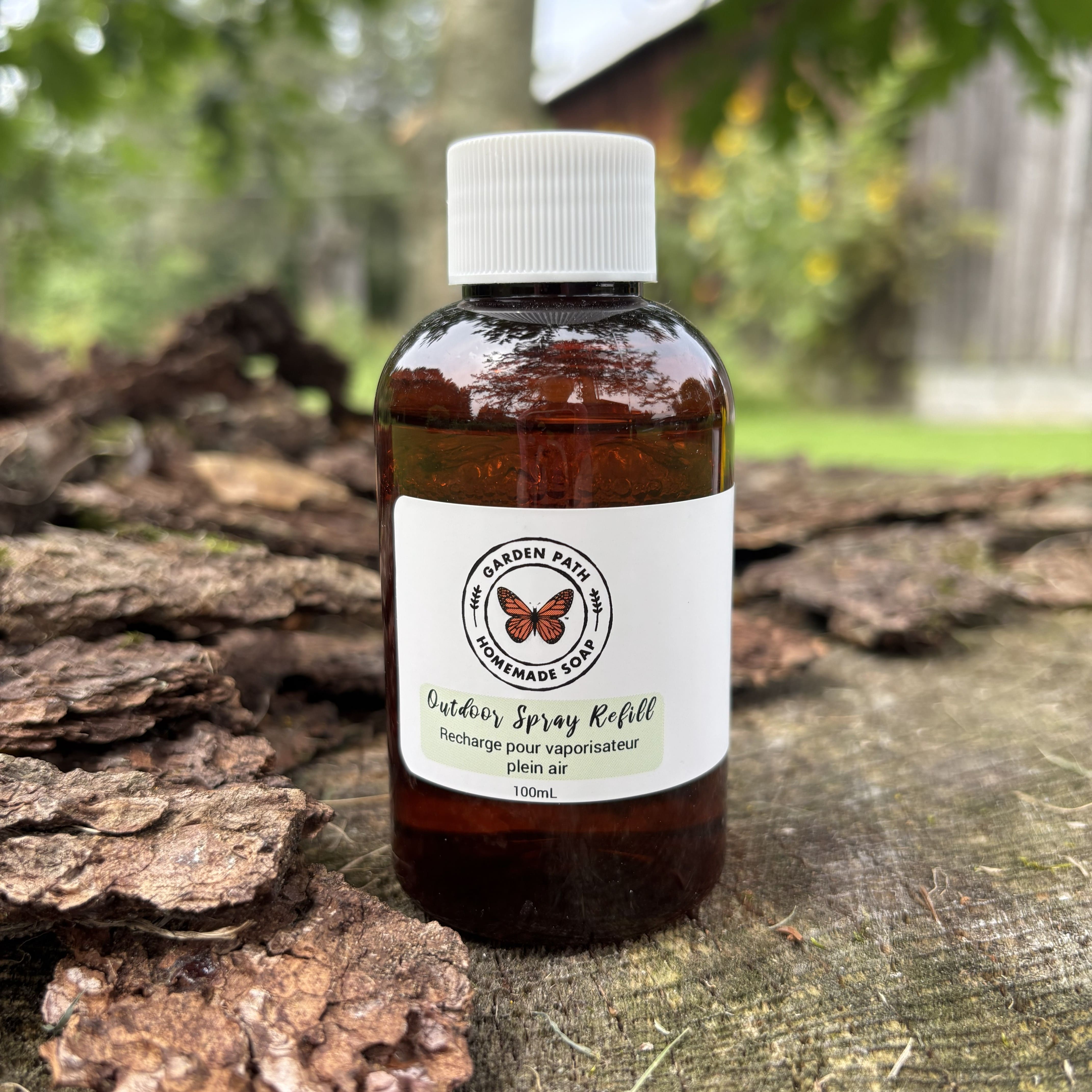 A 100ml amber glass bottle of Outdoor Spray Refill with a white cap and white label, placed on a rustic log with a blurred outdoor garden background. 100% Natural Essential Oil Blend - Garden Path Homemade Soap