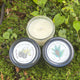 Two candles with labels on a green leafy background. One candle open to see the wick