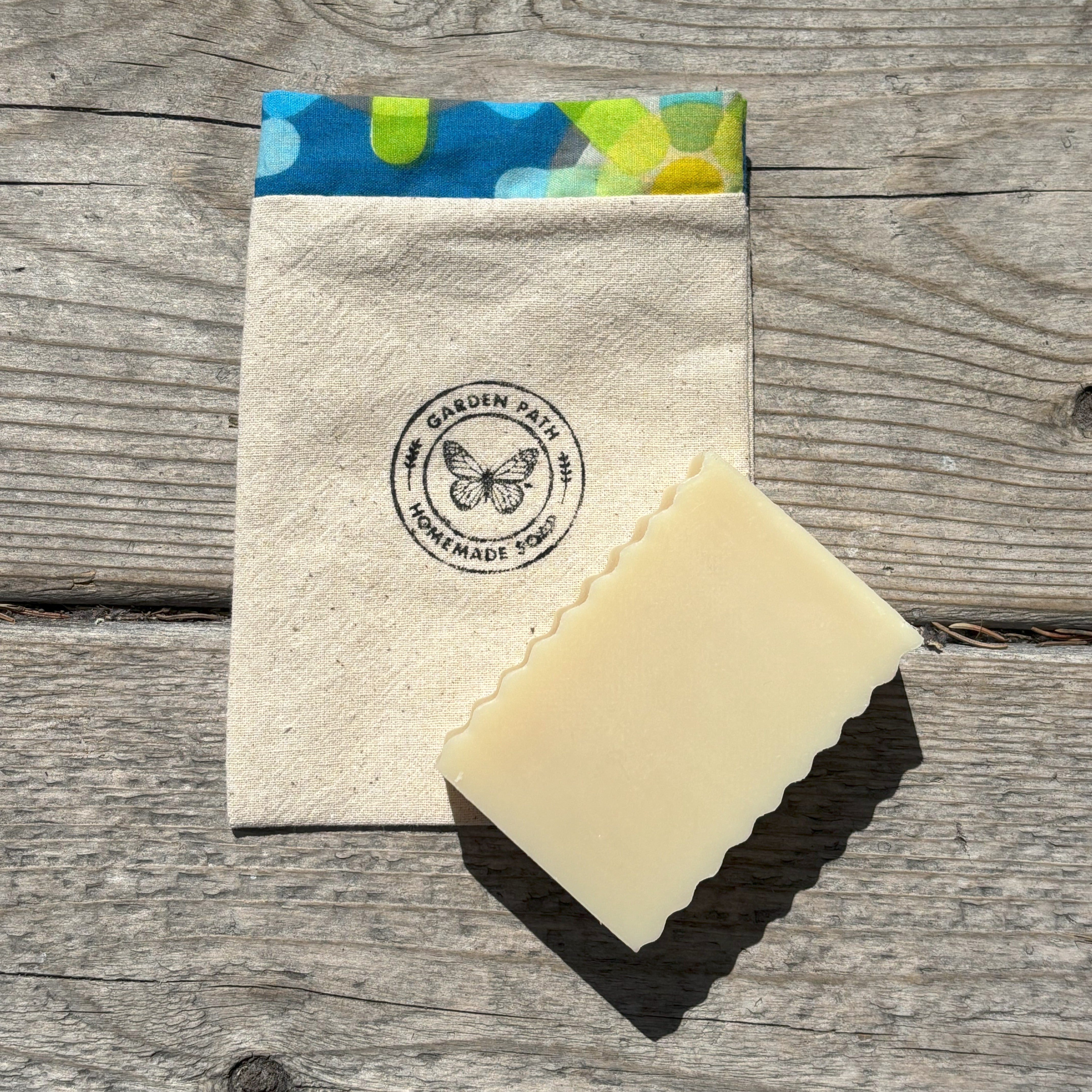 A handmade soap travel bag with a snap closure, featuring a colorful trim and the 'Garden Path Homemade Soap' logo on the front. A bar of soap is placed in front of the bag.