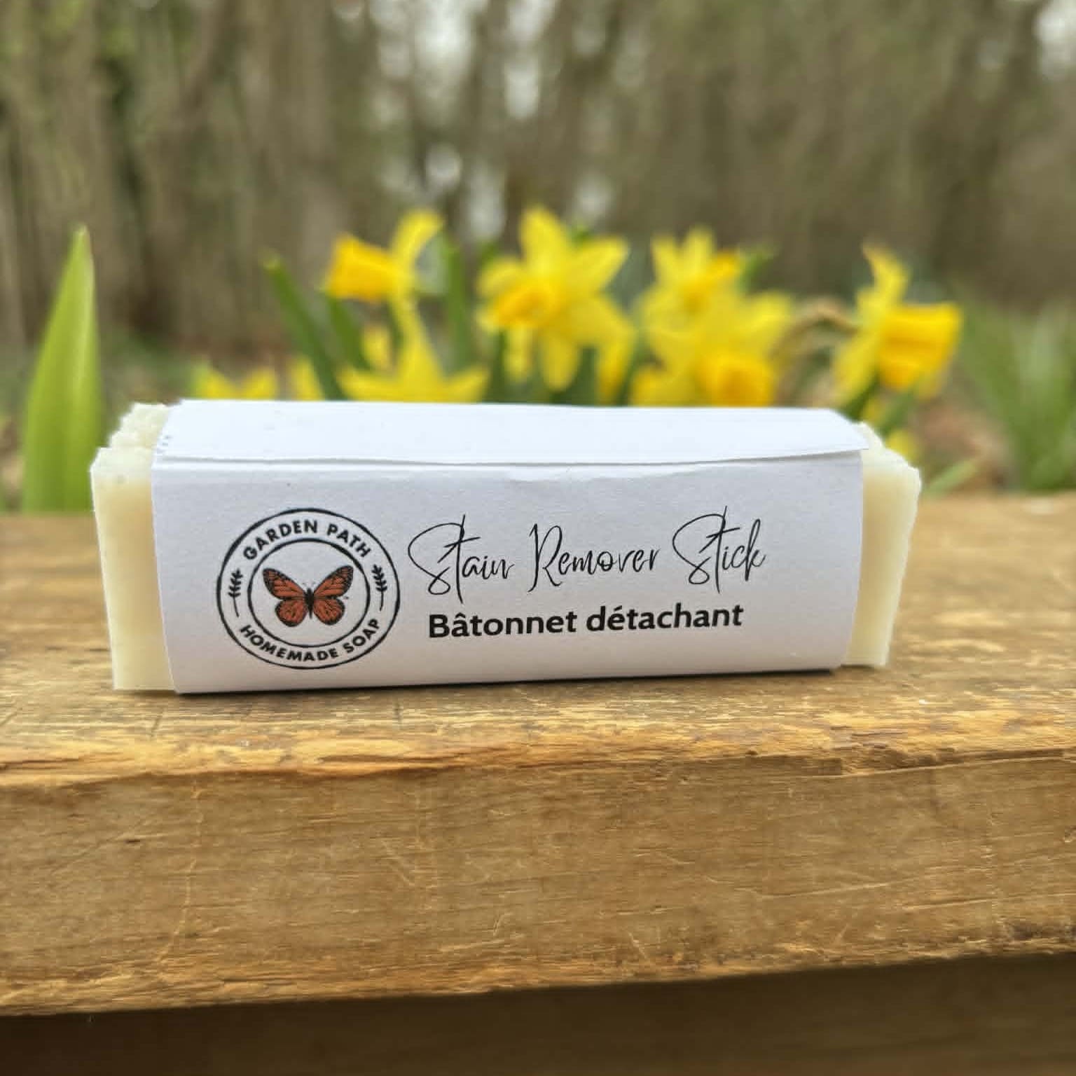 Stain remover stick with a butterfly logo of Garden Path Homemade Soap on a wooden surface with yellow flowers in the background. 100% Natural Ingredients