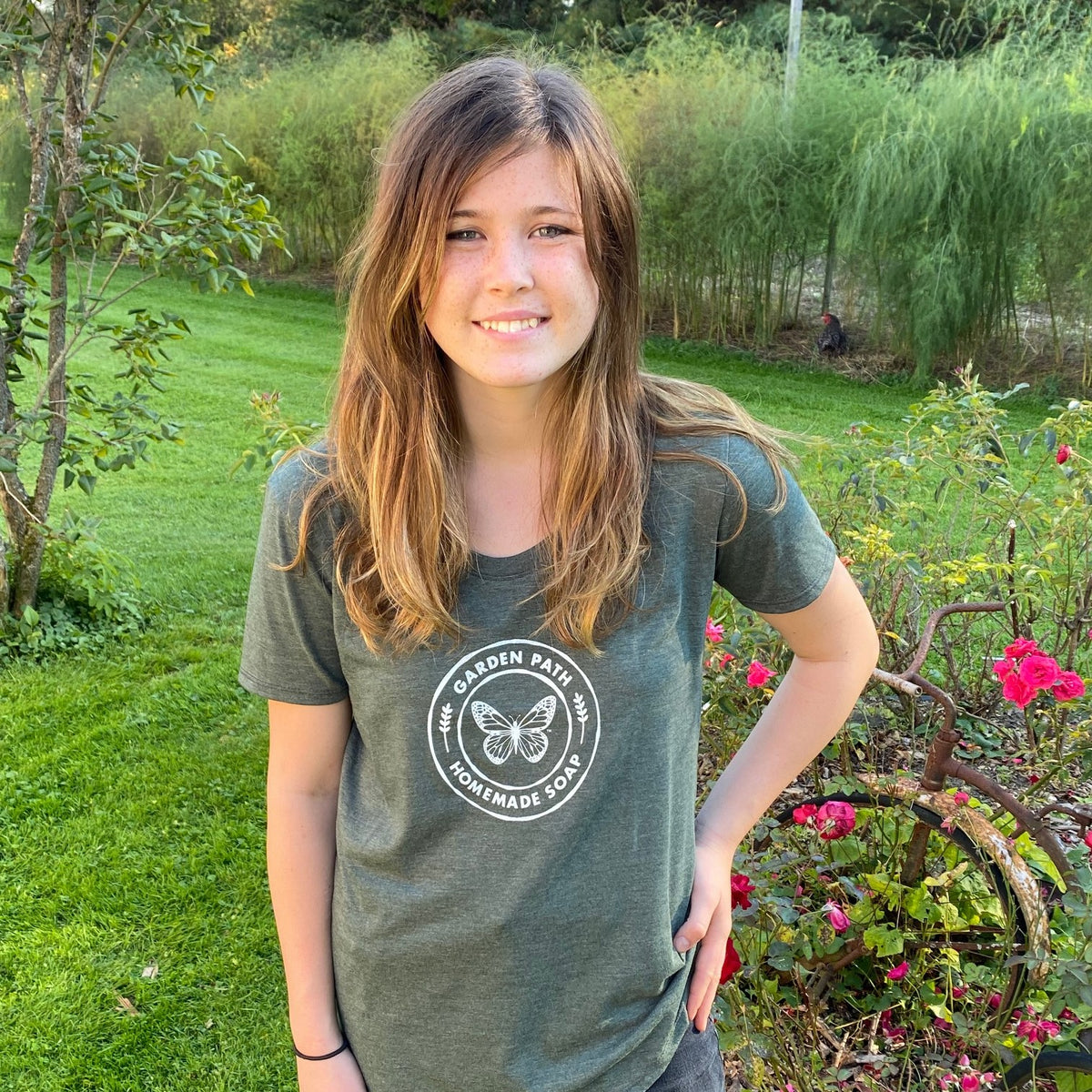 A young girl standing outdoors wearing a green t-shirt with the Garden Path Homemade Soap logo on it.
