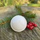 Frosted Winterberry Bath Bomb with a frosted white exterior and red speckles, placed on a wooden surface with green pine needles and red berries to the side.