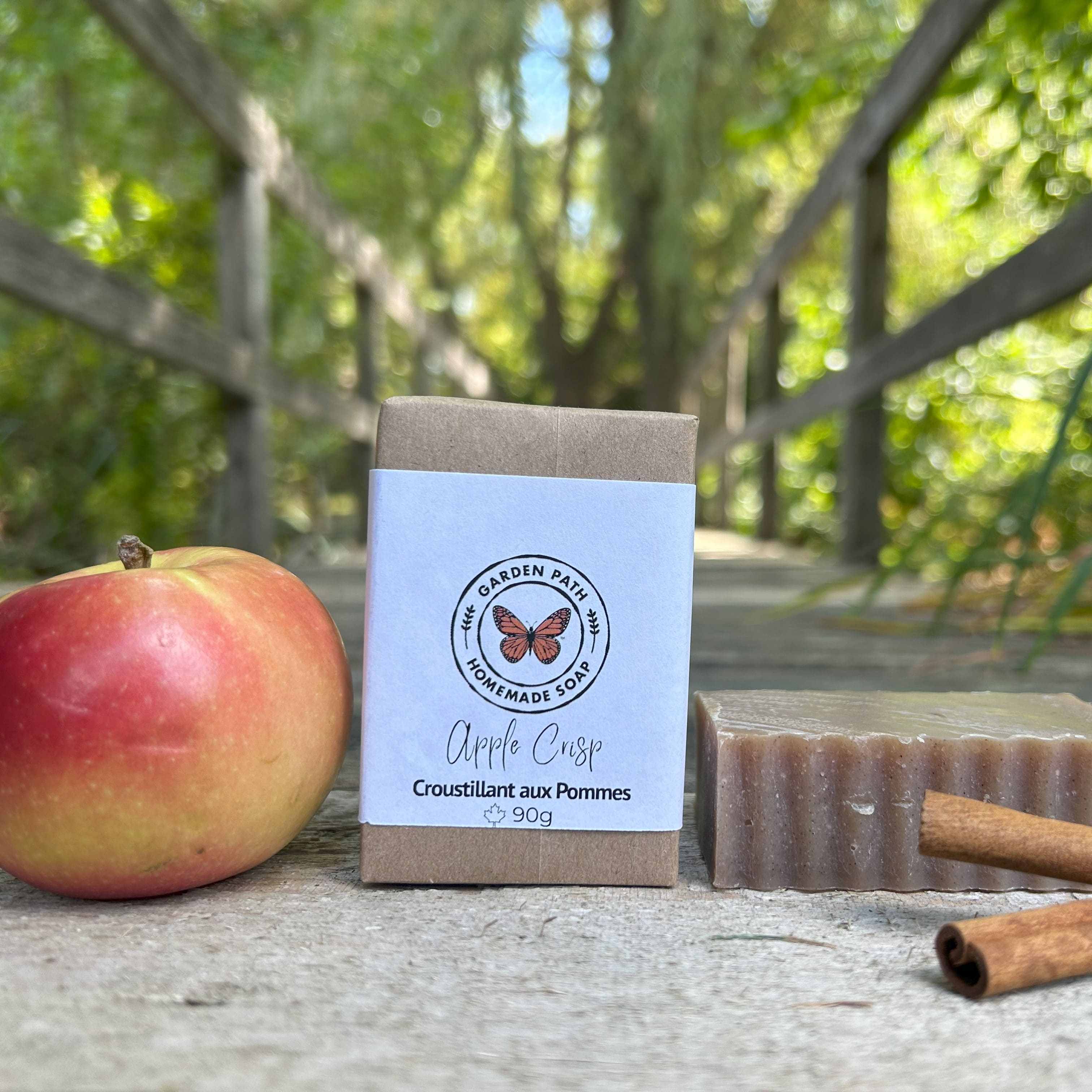 Bar of soap with packaging labeled 'Apple Crisp' next to an apple and cinnamon sticks outdoors by Garden Path Homemade Soap.  Exfoliating with Spicy Fragrance Oils