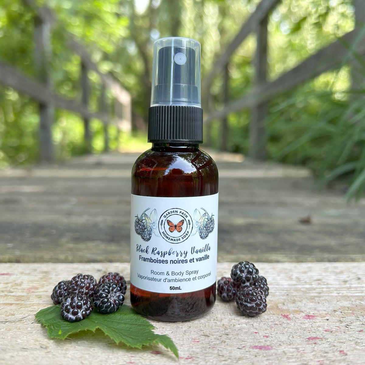 Black Raspberry Vanilla Room and Body Spray bottle by Garden Path Homemade Soap on a wooden path with black raspberries.