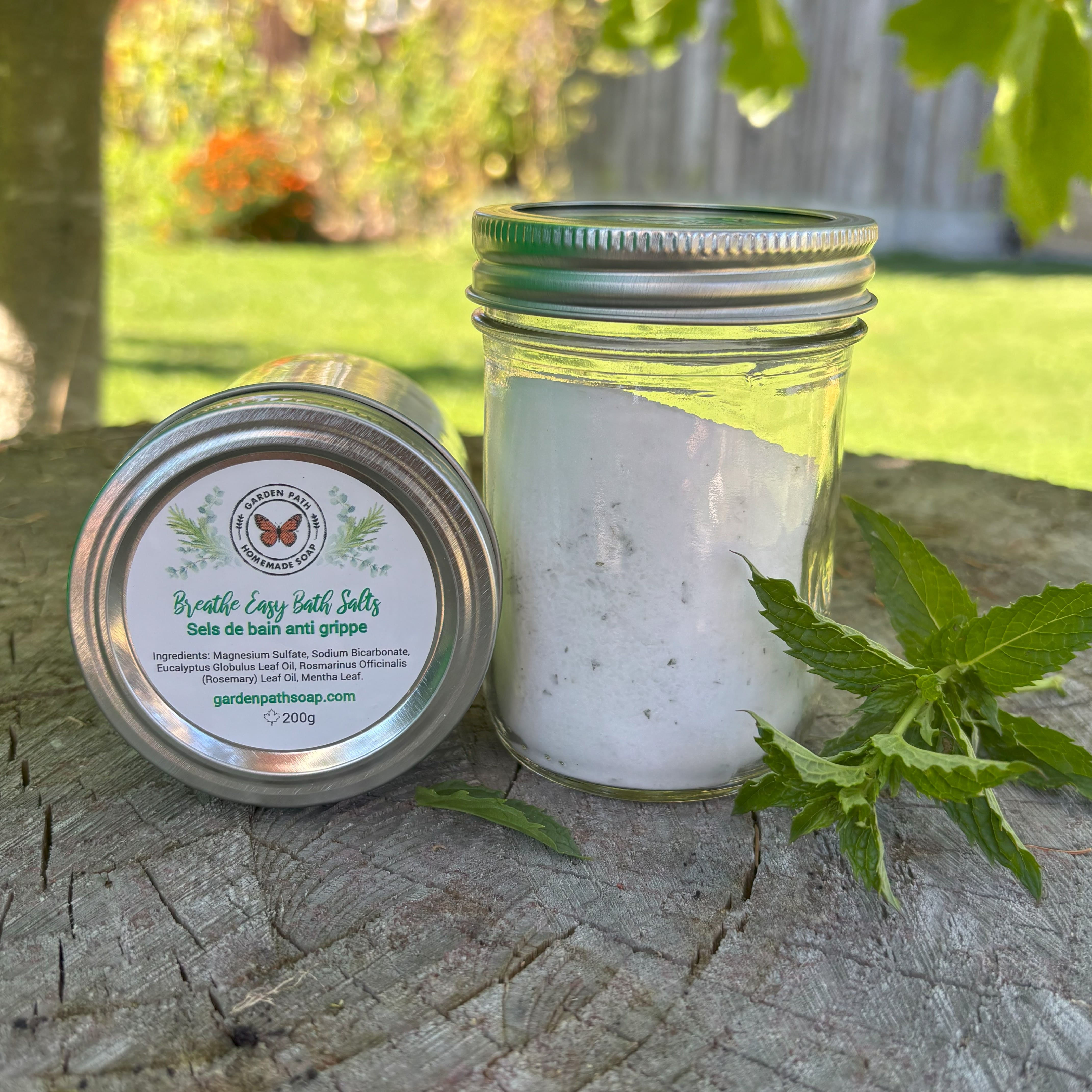 Two containers of 'Breathe Easy Bath Salts' on a wooden surface with greenery in the background and a sprig of mint lying beside the bottles by Garden Path Homemade Soap. 100% Natural Essential Oils & Ingredients