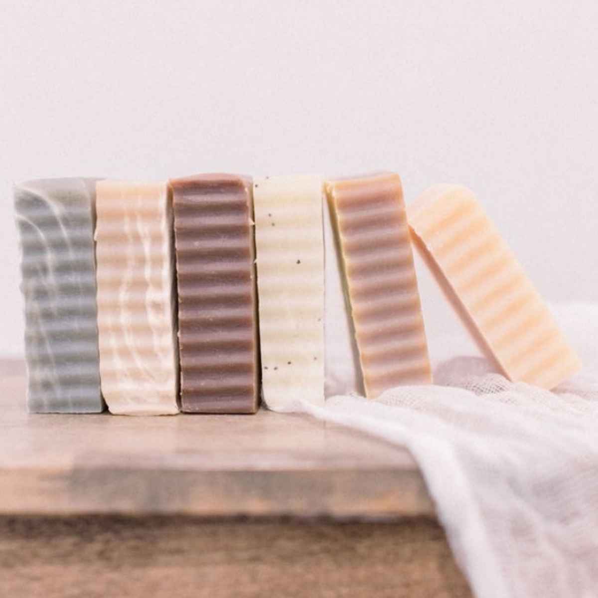 Six rectangular handmade soap bars in assorted neutral and earthy colors, arranged upright on a wooden surface with a white cloth draped behind them.