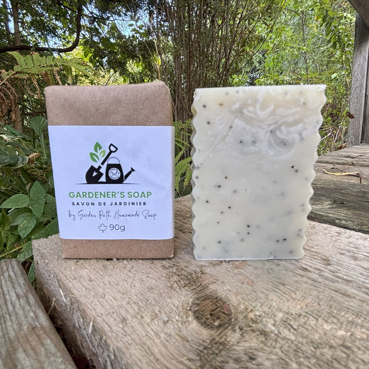 Gardener's Soap Bar on a wooden surface. 2 bars-one wrapped and one unwrapped. Label show shovel and watering can with Garden Path Homemade Soap Label.