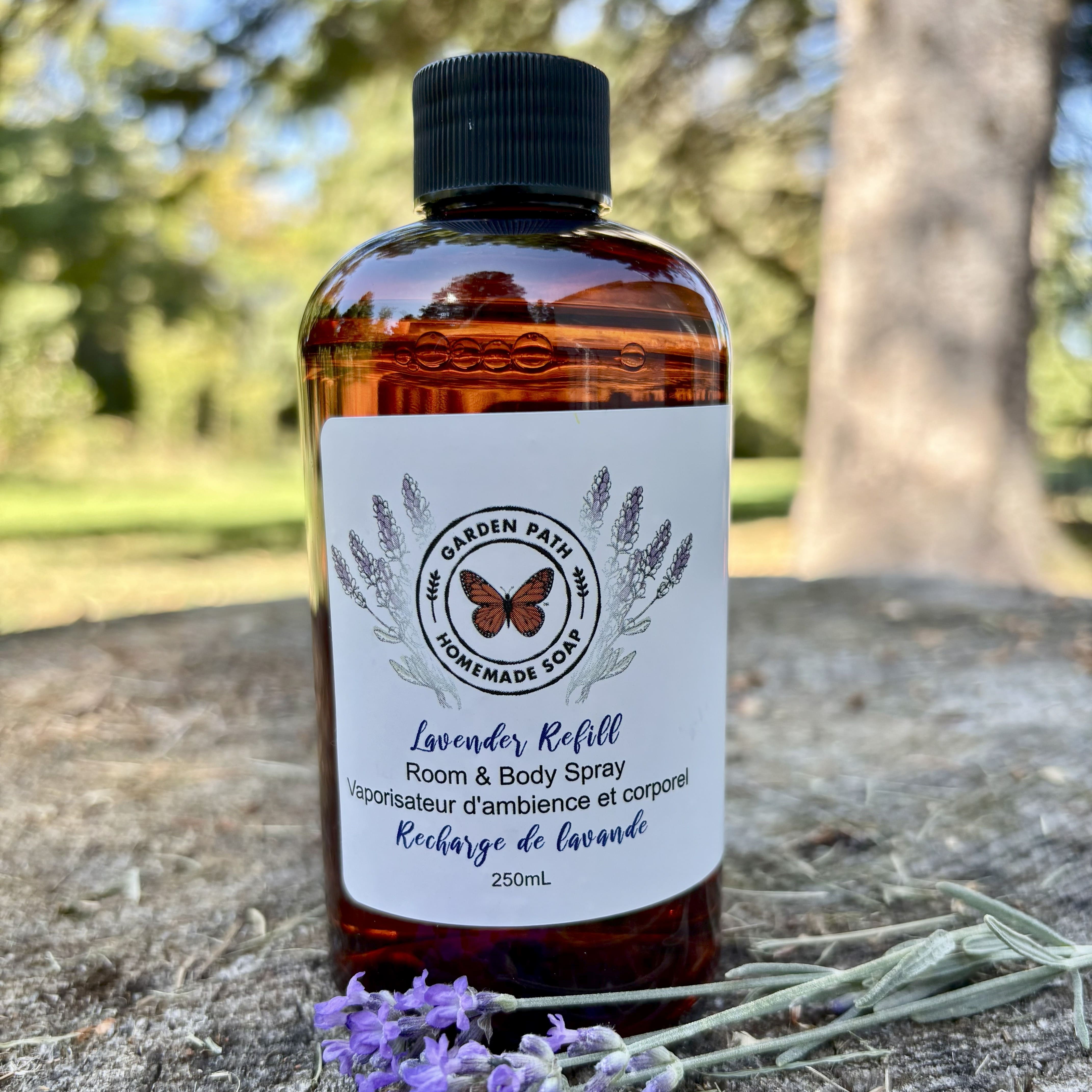 Bottle of lavender refill room and body spray sitting on a wooden surface with lavender flowers by Garden path Homemade Soap.  100% Natural Essential Oil