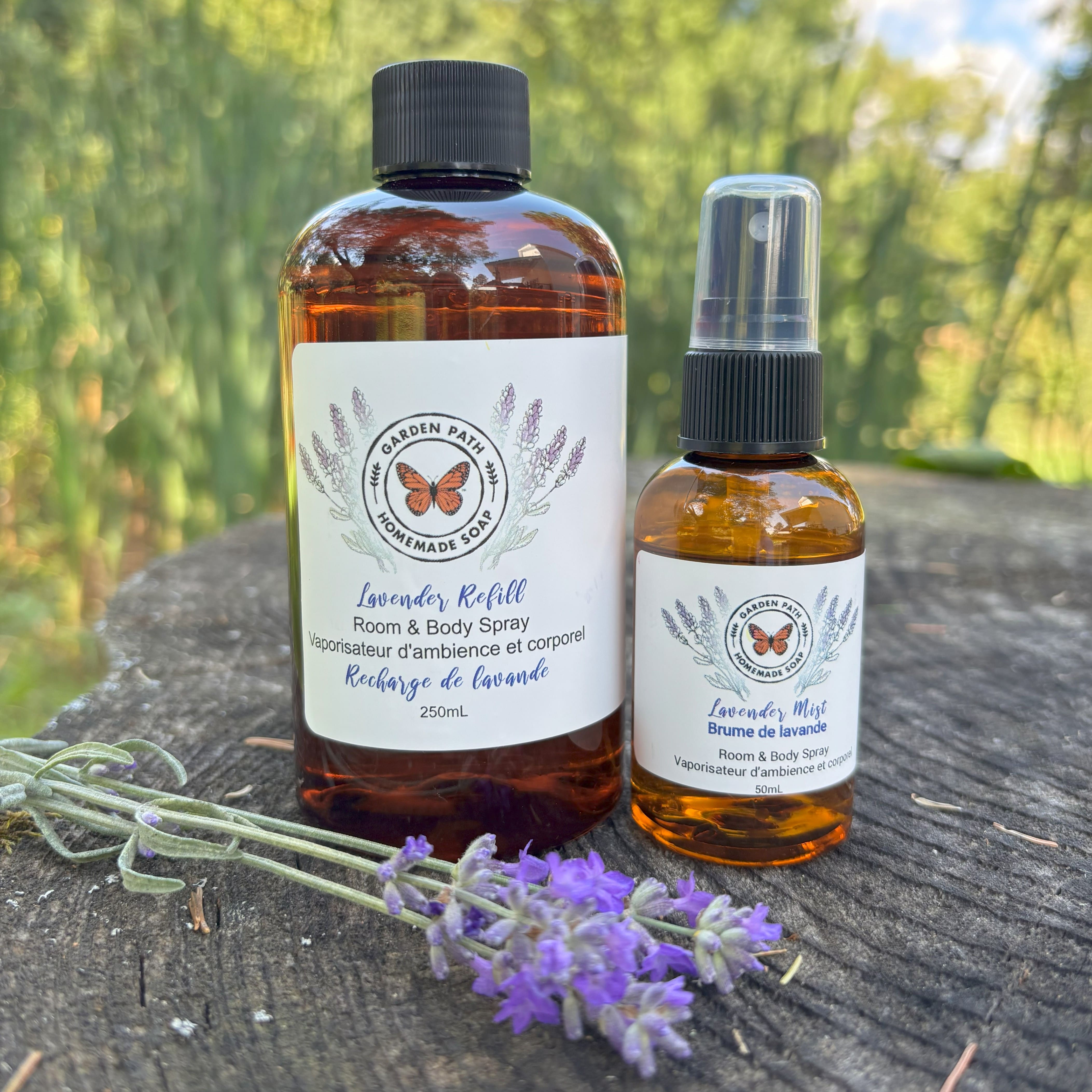 Two bottles of lavender-scented room, one a spray bottle and one a refill bottle  on a wooden surface with lavender flower by Garden Path Homemade Soap.  100% Natural Essential Oil