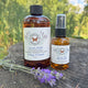 Two bottles of lavender-scented room, one a spray bottle and one a refill bottle  on a wooden surface with lavender flower by Garden Path Homemade Soap.  100% Natural Essential Oil