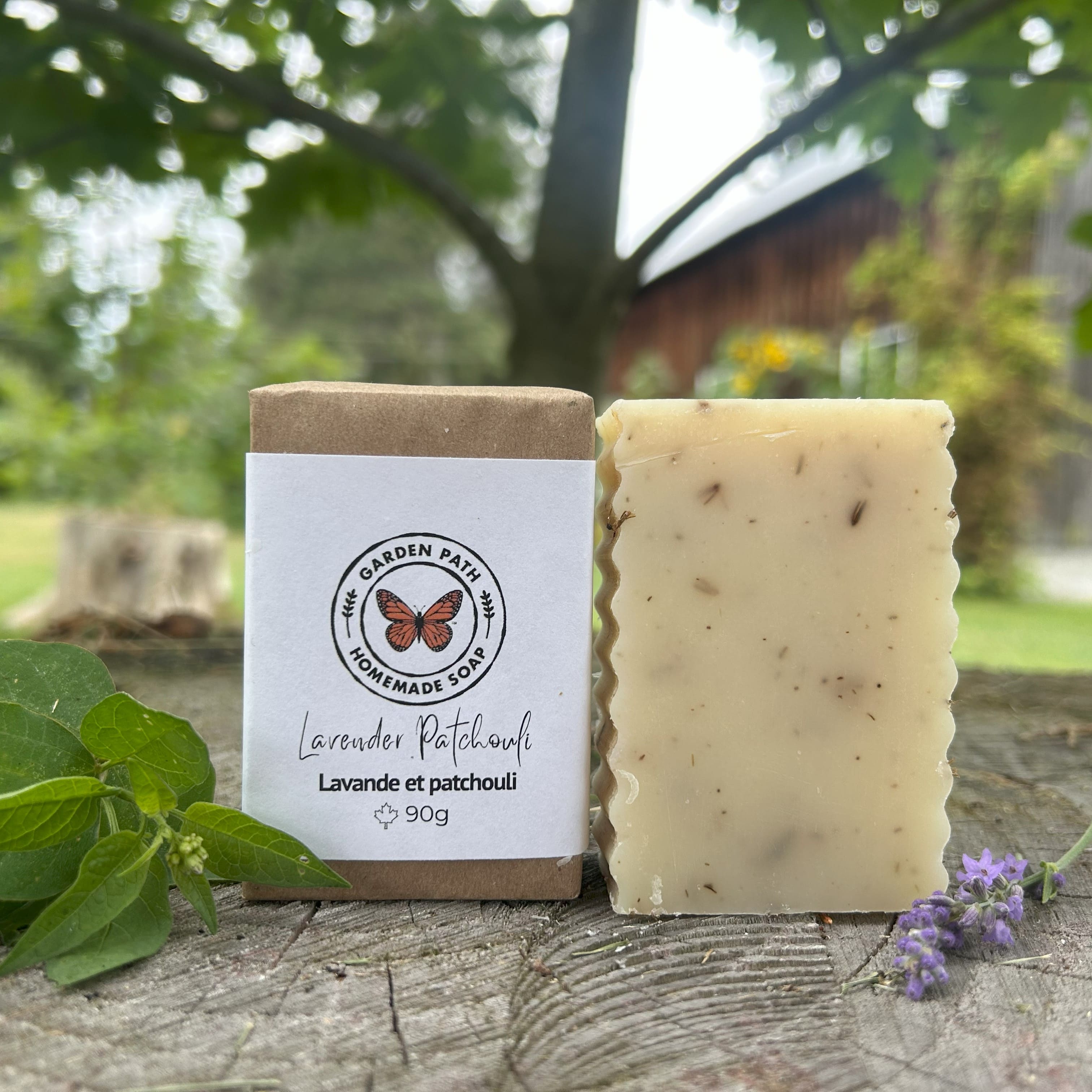 Lavender Patchouli Bar of soap with a label on a wooden surface outdoors. One bar wrapped the other unwrapped.  Made with 100% Natural Essential Oils and Lavender Buds  by Garden Path Homemade Soap