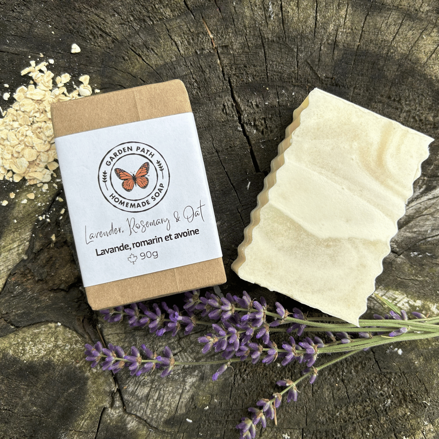 A 90g bar of handmade soap by Garden Path Homemade soap  labeled 'Lavender, Rosemary & Oat' sits next to its kraft paper packaging on a rustic wood surface, styled with dried lavender flowers and oats.