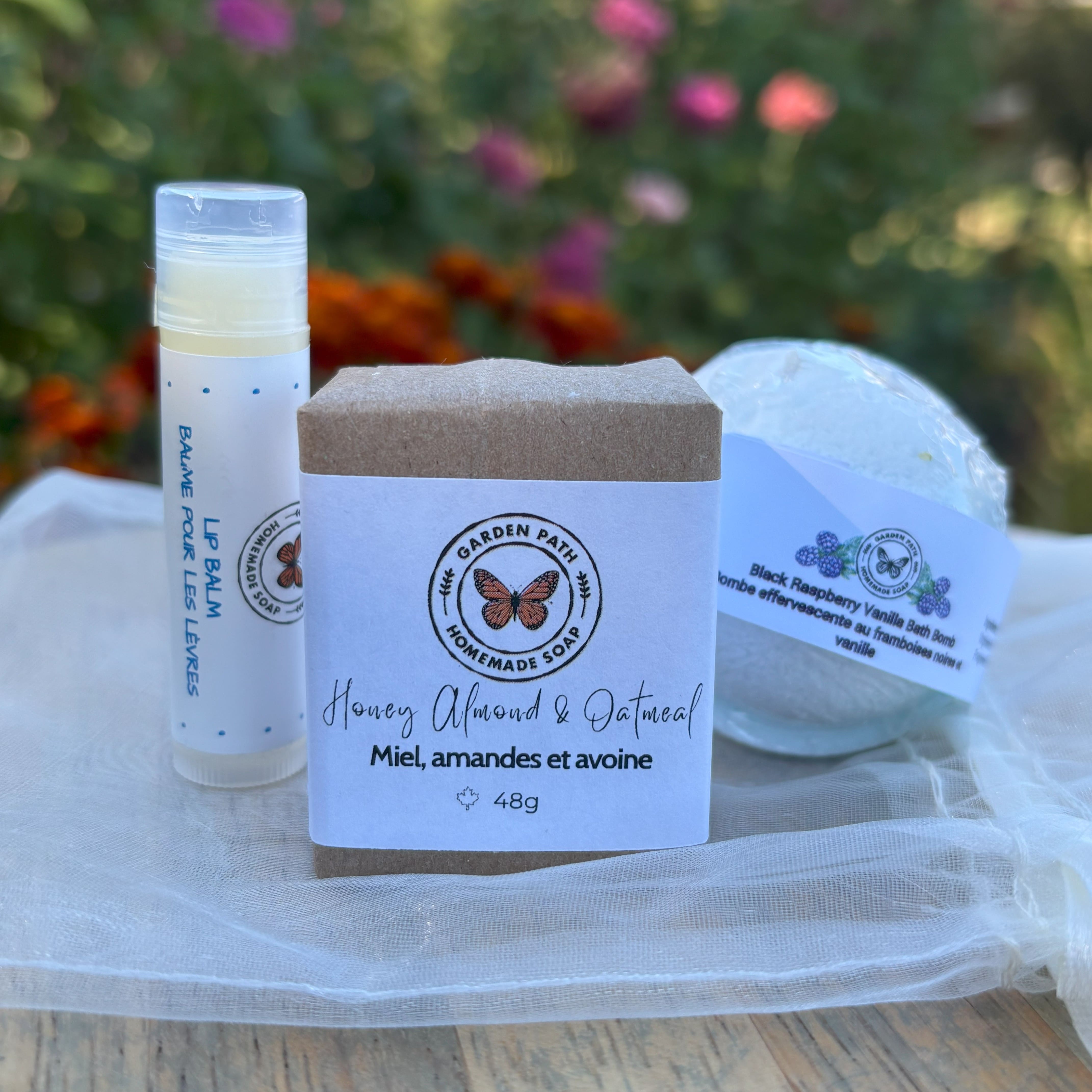 Lip balm, soap bar, and bath bomb from Garden Path Homemade Soap on a white organza fabric with a blurred floral background by Garden Path Homemade Soap.