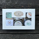Spa in a Box set with various products like bar soap, facial scrub, small candle, shower puck and lip balm on a wooden surface by Garden Path Homemade Soap