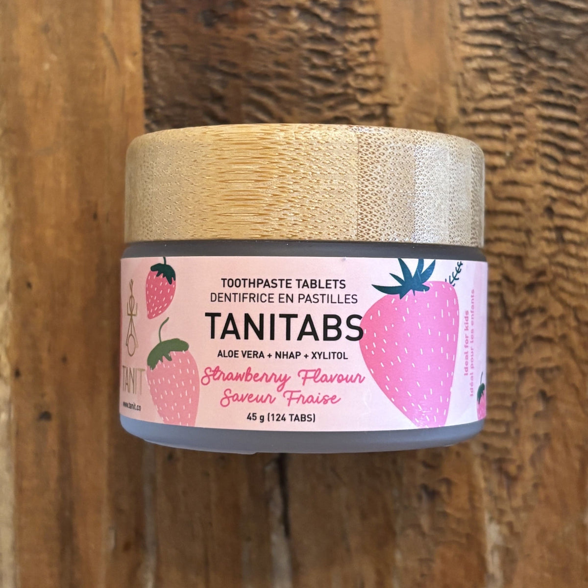 A jar of TANITABS Strawberry Flavour toothpaste tablets, featuring a light wood lid and a pink label decorated with illustrations of strawberries, set against a rustic wooden background.