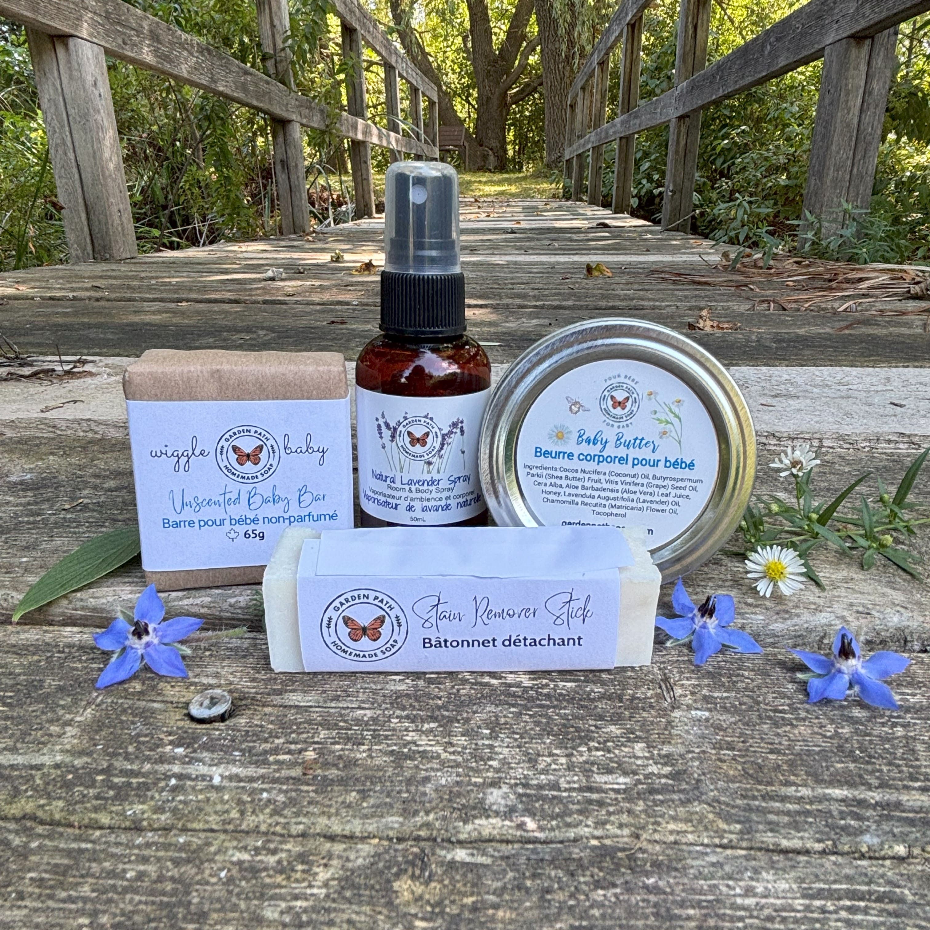 Wiggle Baby Bundle by Garden Path Homemade Soap with wiggle bar, lavender spray, baby butter, stain remover stick on a wooden surface with  some blue tiny flowers.