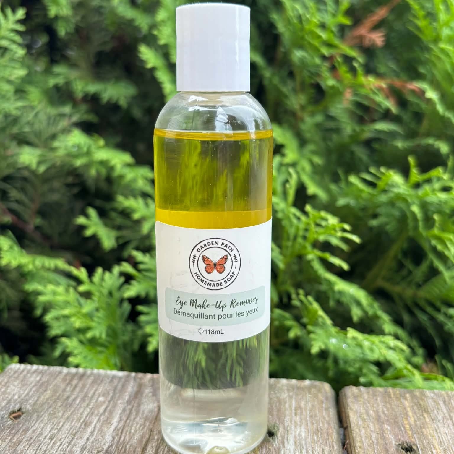 Eye make up remover bottle sitting on a wooden surface with cedar tress in the background by Garden Path Homemade Soap. 100% Natural Ingredients