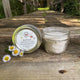 Jar of face scrub with a metal lid on a wooden surface with a natural background by Garden Path Homemade Soap. 100% Natural Ingredients