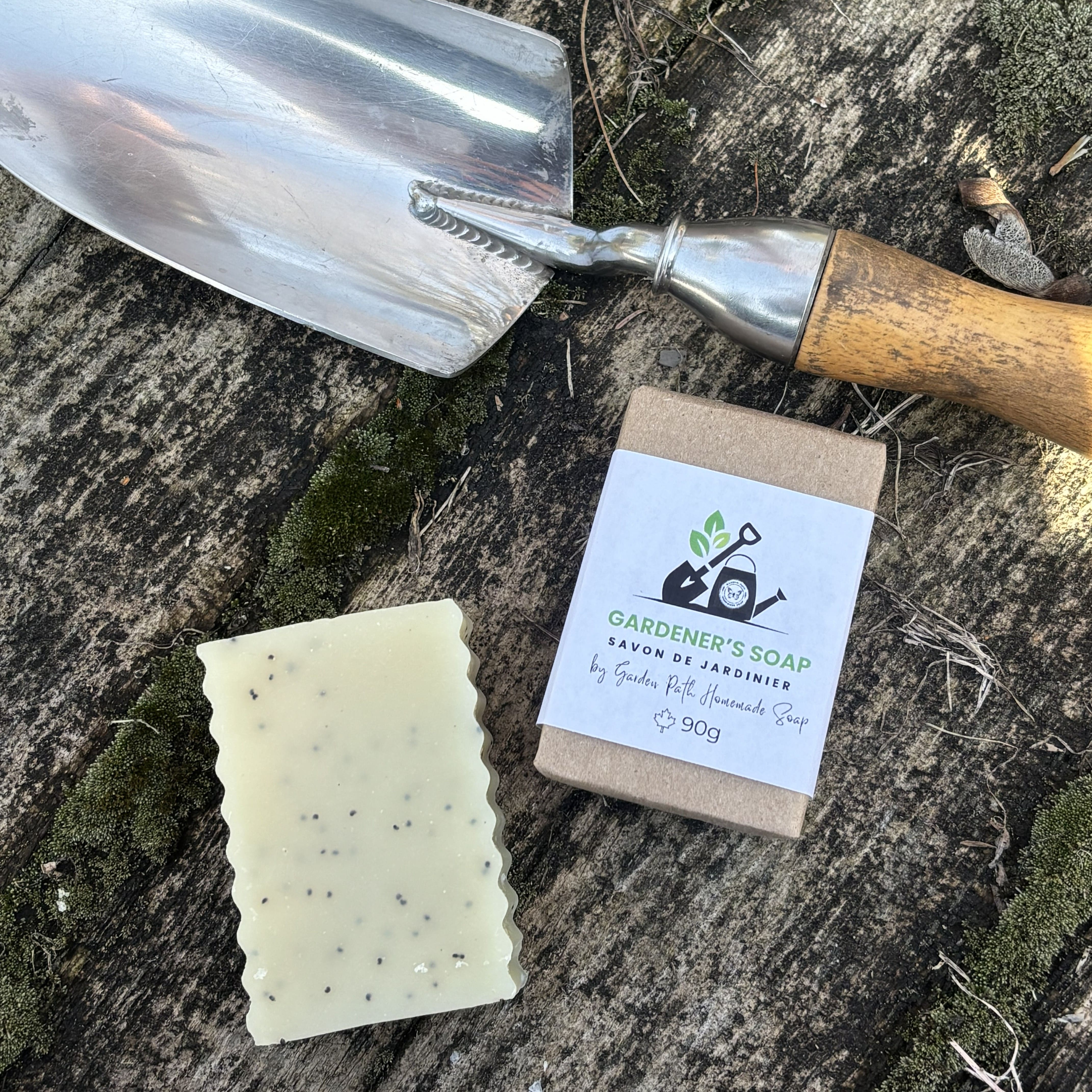Gardener's Soap Bar on a wooden surface. 2 bars-one wrapped and one unwrapped. Label show shovel and watering can with Garden Path Homemade Soap Label. 