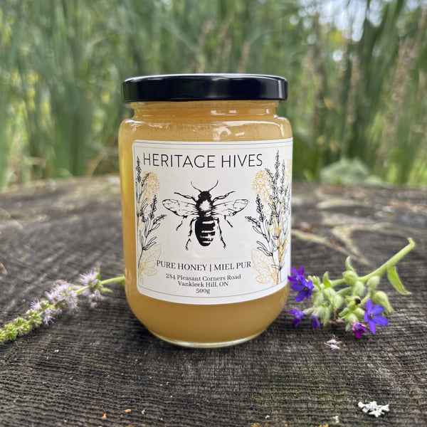A jar of Heritage Hives Pure Honey  with a black lid and white label, set on a dark wooden surface outdoors with green foliage in the background and small purple flowers in the foreground.