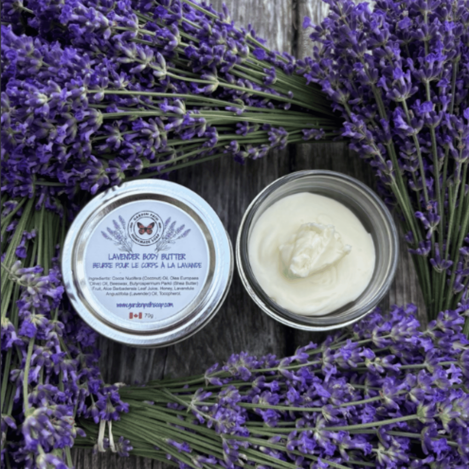 Lavender body butter in a clear round jar with a white lid labeled 'Lavender Body Butter' by  Garden Path Homemade soap, placed on a rustic wooden surface surrounded by fresh purple lavender sprigs.