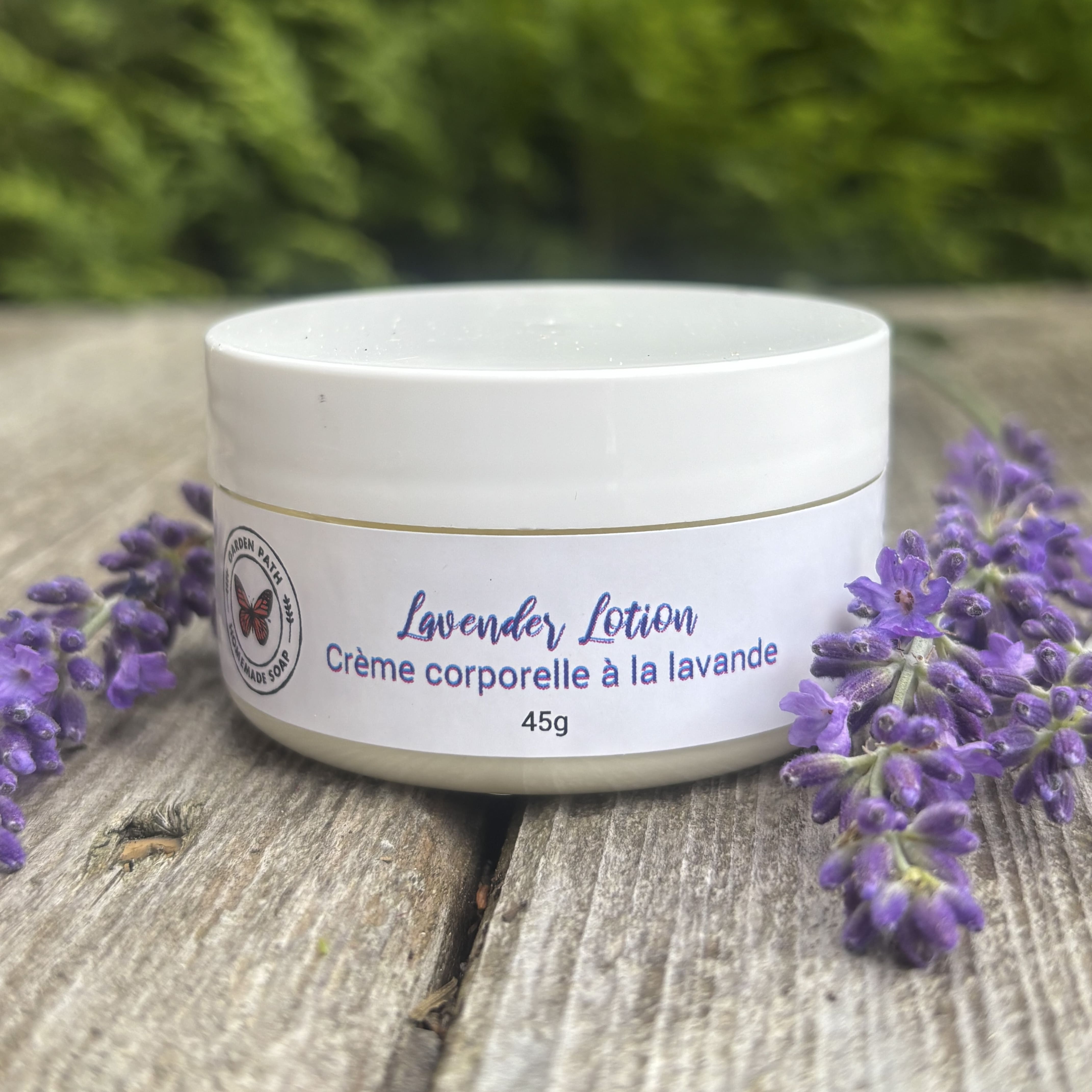 White lavender lotion jar surrounded by purple lavender flowers on wood. 100% Natural Ingredients & Essential Oil by Garden Path Homemade Soap