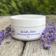 White lavender lotion jar surrounded by purple lavender flowers on wood. 100% Natural Ingredients & Essential Oil by Garden Path Homemade Soap