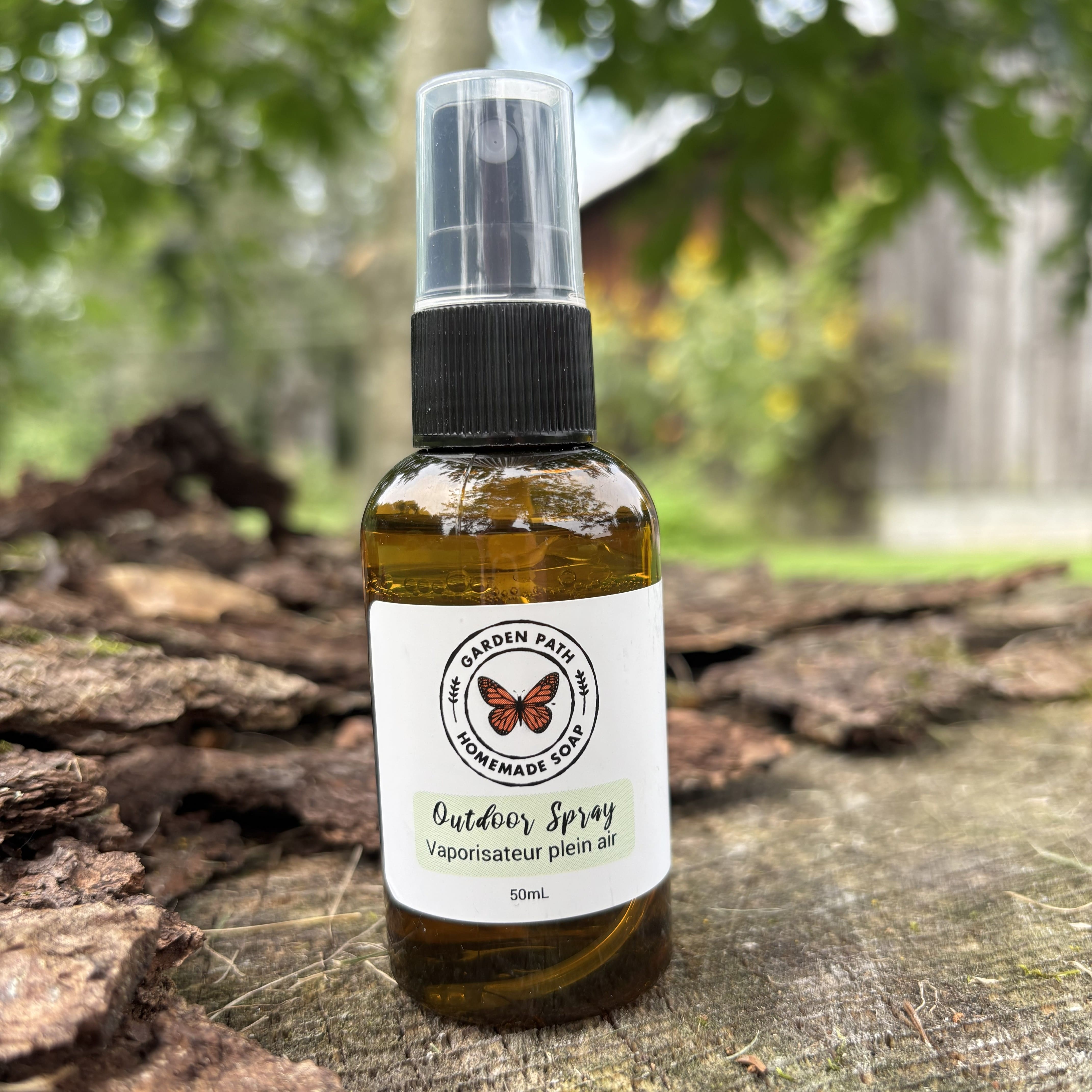 A 50mL amber glass spray bottle of Garden Path Outdoor Spray with a black nozzle and clear cap, resting on a rustic tree trunk with a blurred outdoor garden background.  100% Natural Essential Oil Blend - Garden Path Homemade Soap