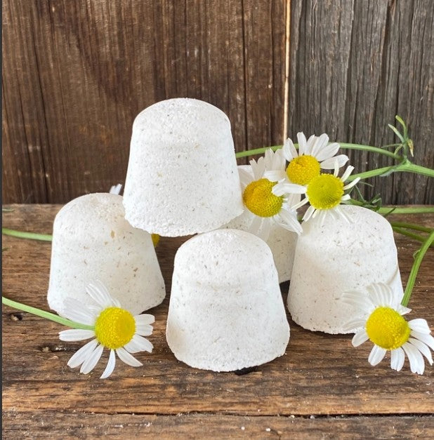 Four white shower tablets arranged on a rustic wooden surface with white daisies, set against a dark wood plank background. 100% Natural Essential Oils Aromatherapy by Garden Path Homemade Soap