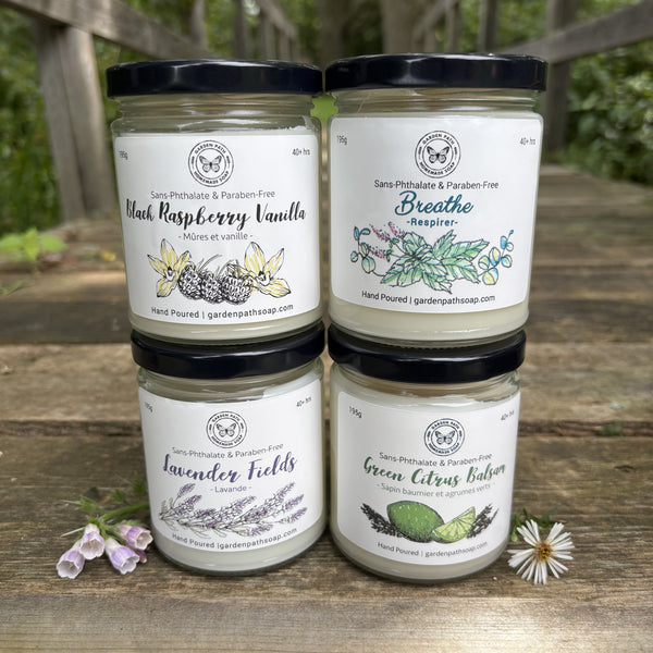 Four large  soy candles  arranged in a 2x2 grid on a rustic wooden table. The background shows a blurred outdoor garden setting.  Garden Path Homemade Soap