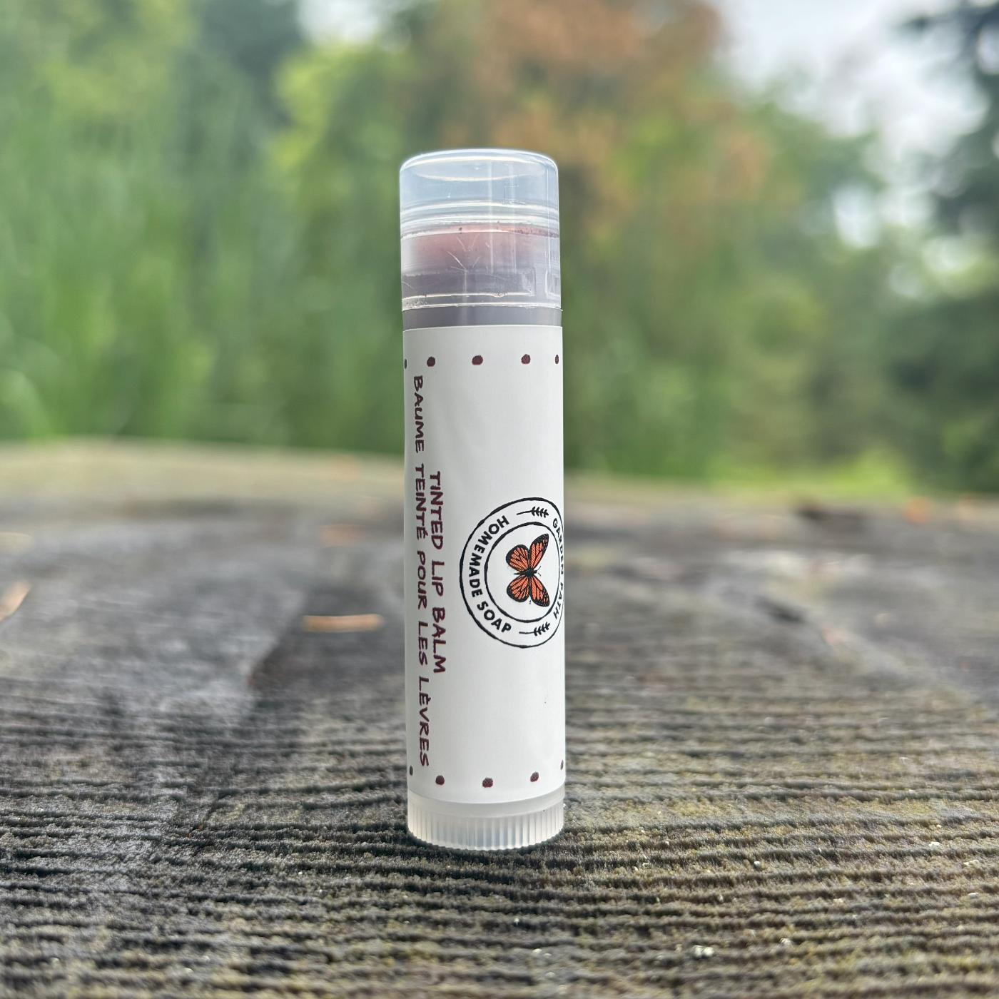 A stick of tinted lip balm with 100% natural ingredients displayed on a wooden surface by Garden Path Homemade Soap.
