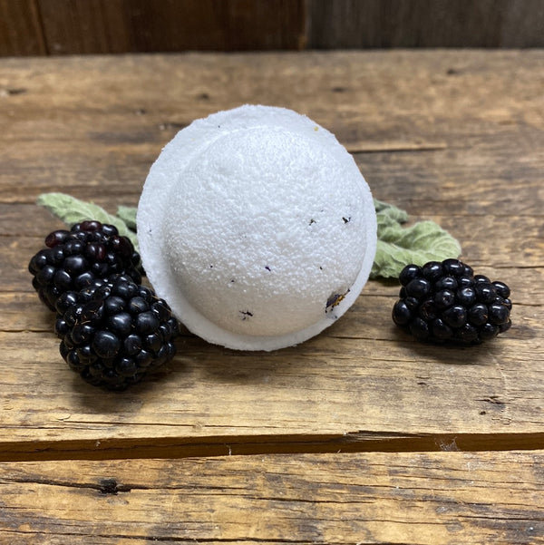A bath bomb with a smooth, white exterior  placed on a wooden surface with black raspberries beside it.