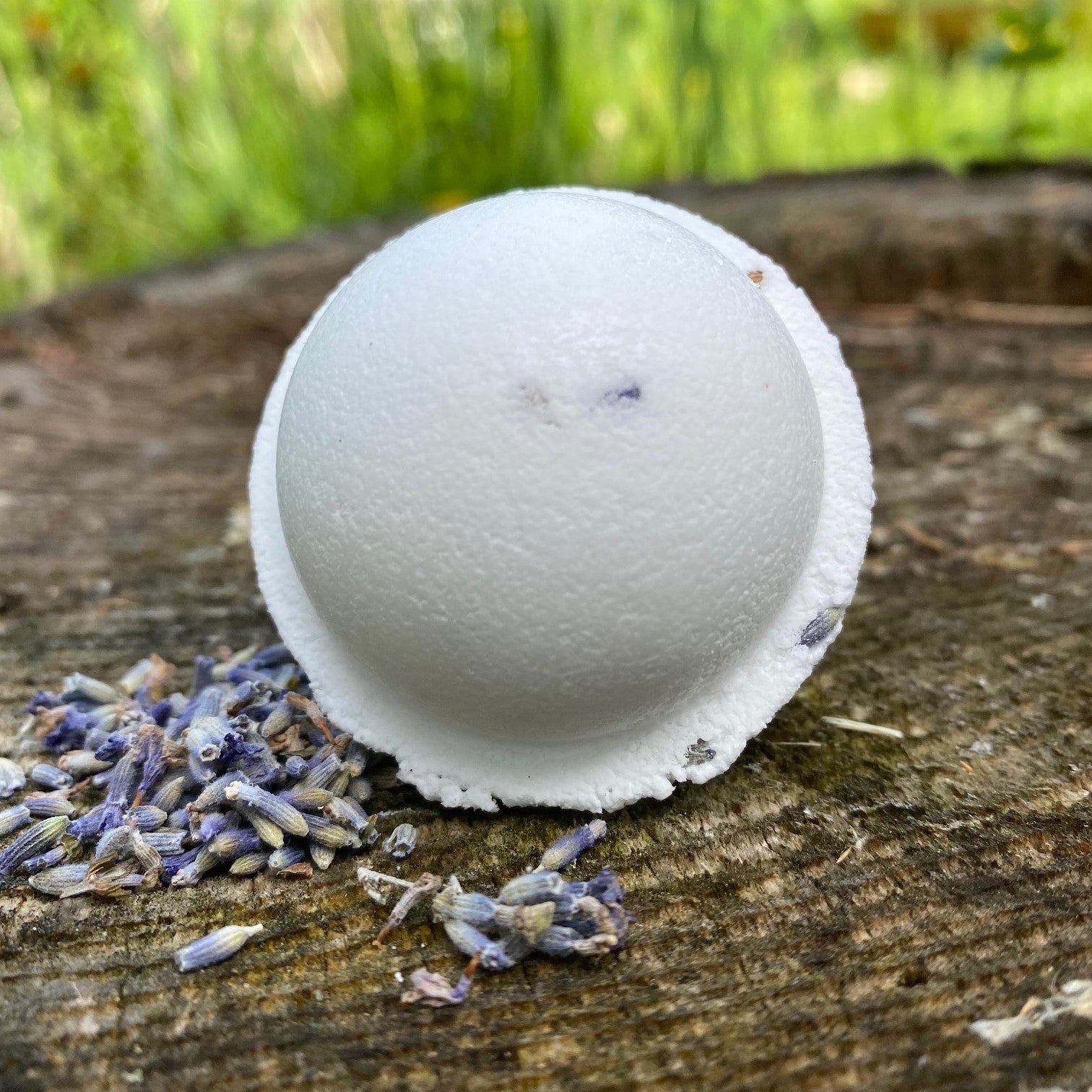 A white lavender  bath bomb with lavender buds visible on the surface, placed on a wooden surface with a scattering of lavender buds around it by Garden Path Homemade Soap. 100% Natural Essential Oil & Ingredients