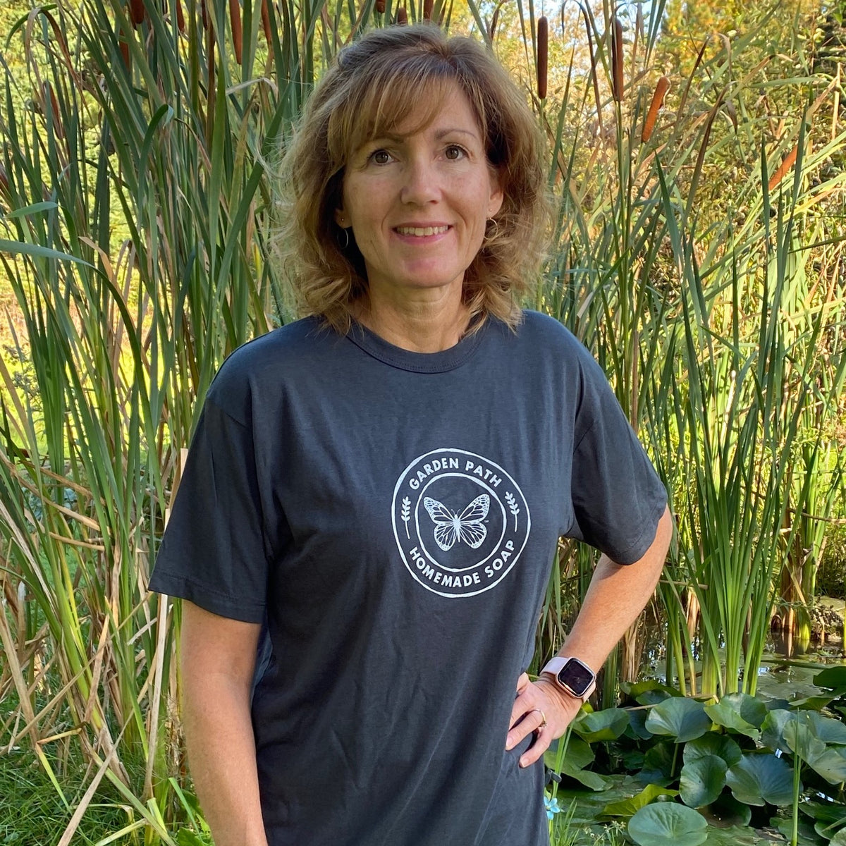 A woman wearing a bamboo cotton t-shirt with the Garden Path Homemade Soap logo on it. T-shirt is charcoal grey